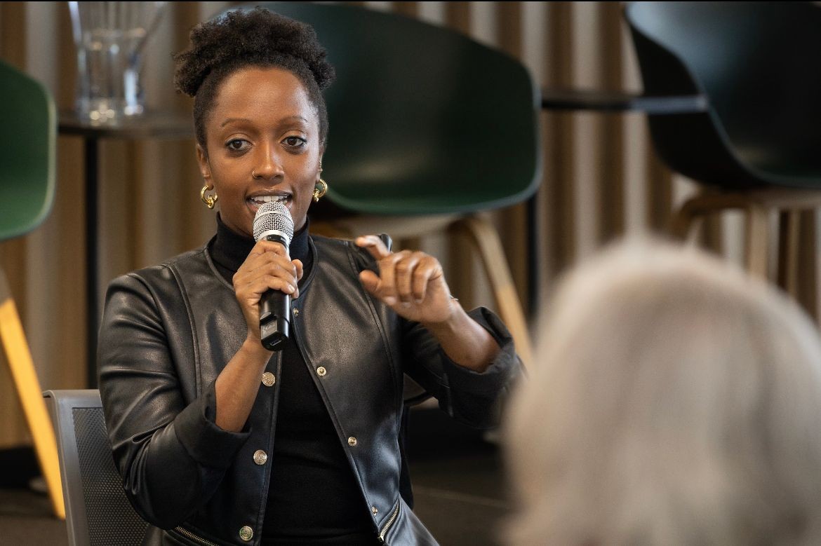 Junie in a black leather jacket holding a microphone speaking at Women in Emerging Tech to a diverse audience of tech companies and tech professionals on inclusive talent in tech.